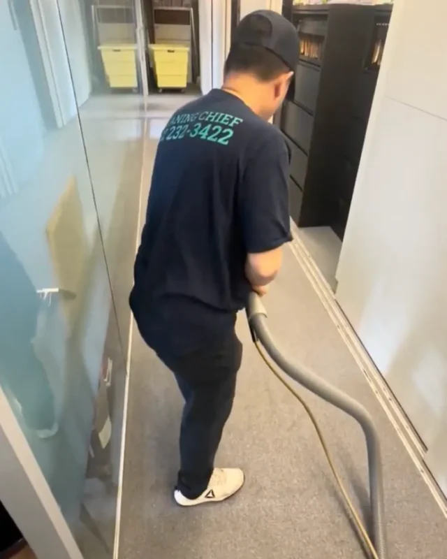 Cleaning Chief technician crouching with carpet extractor hose along base of glass partition wall in commercial office corridor