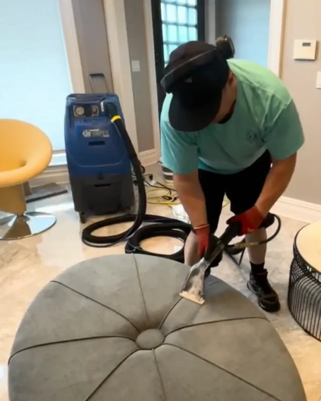 Cleaner using hand-held extractor wand on large round tufted ottoman in commercial lobby, blue carpet machine in background