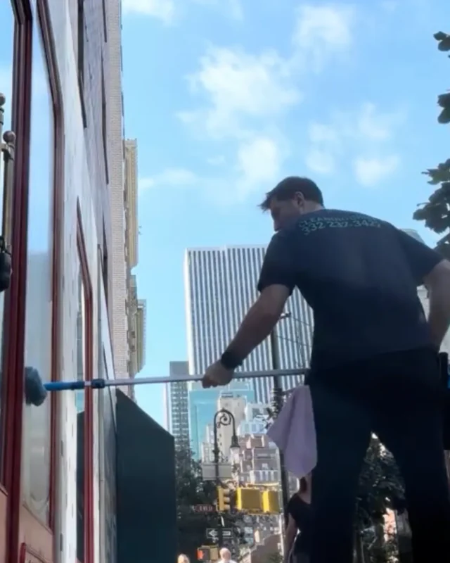 Cleaning Chief cleaner using long extension pole on storefront windows from sidewalk with NYC skyline in background
