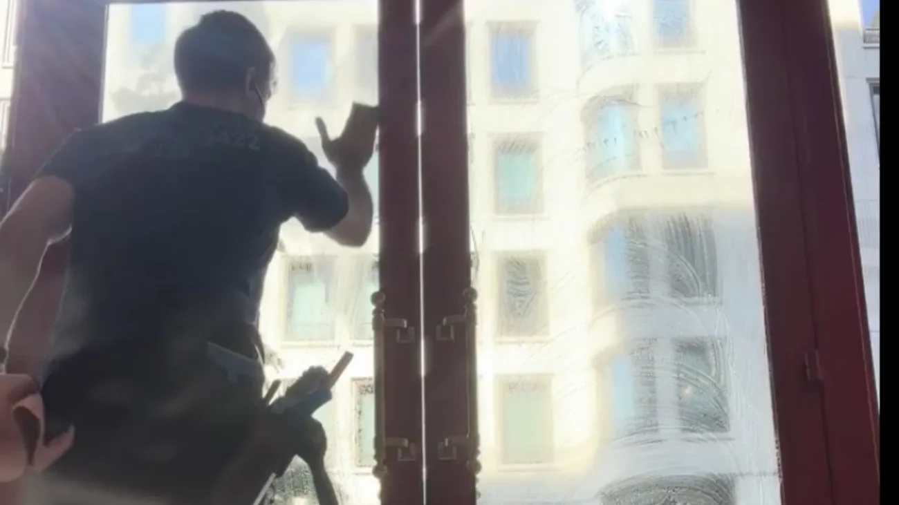 Cleaning Chief cleaner wiping tall red-framed glass doors from inside with ornate NYC building visible through the glass