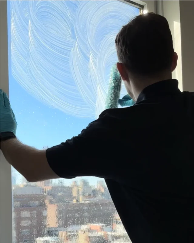 Cleaner scrubbing large interior window with NYC rooftops and buildings clearly visible below