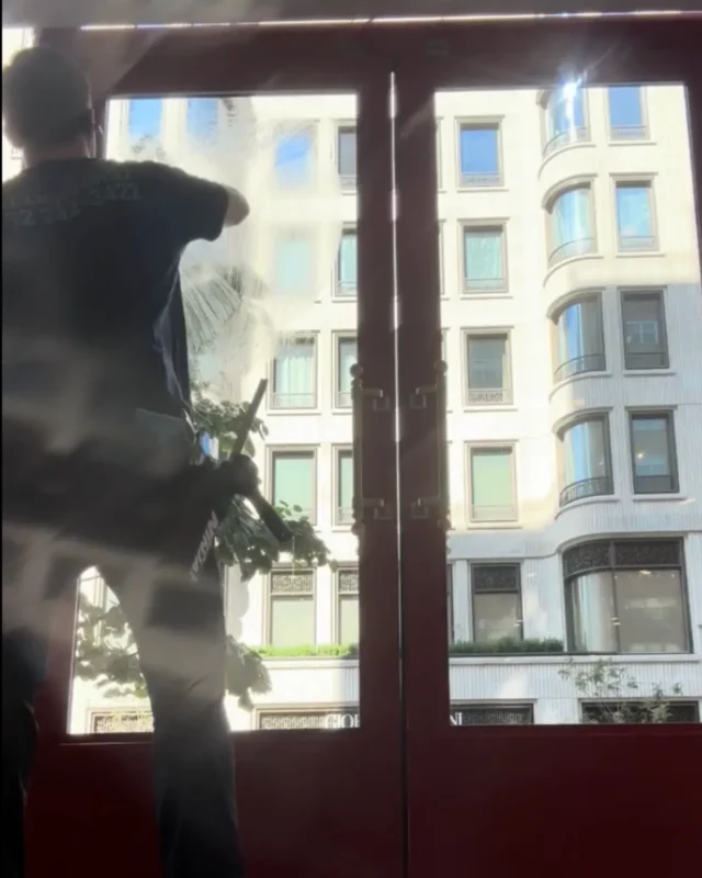Technician silhouetted cleaning tall red-framed glass entry doors with Manhattan building visible through the windows