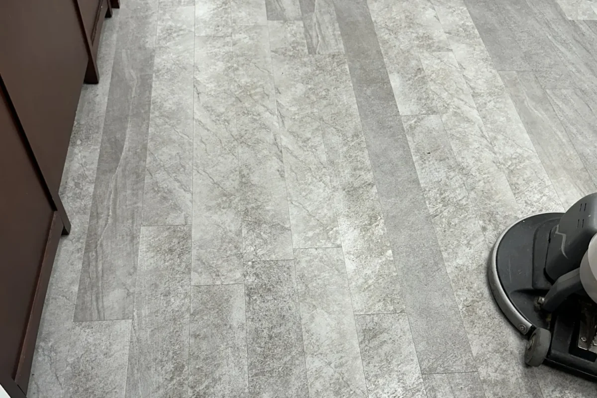 Large-format gray stone tile floor with dull surface and visible wear marks before floor care treatment, buffer at edge