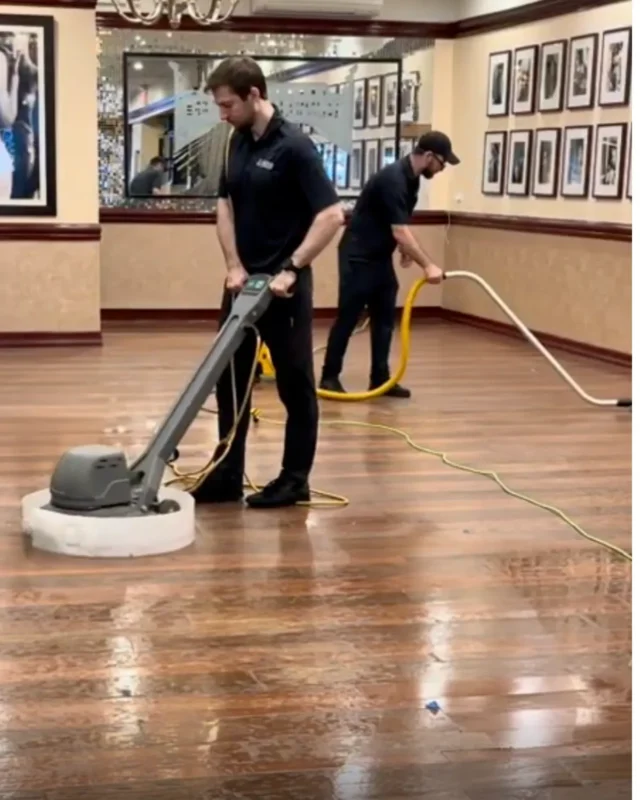Two Cleaning Chief technicians operating floor buffing machine on hardwood floor in NYC commercial hall with framed art