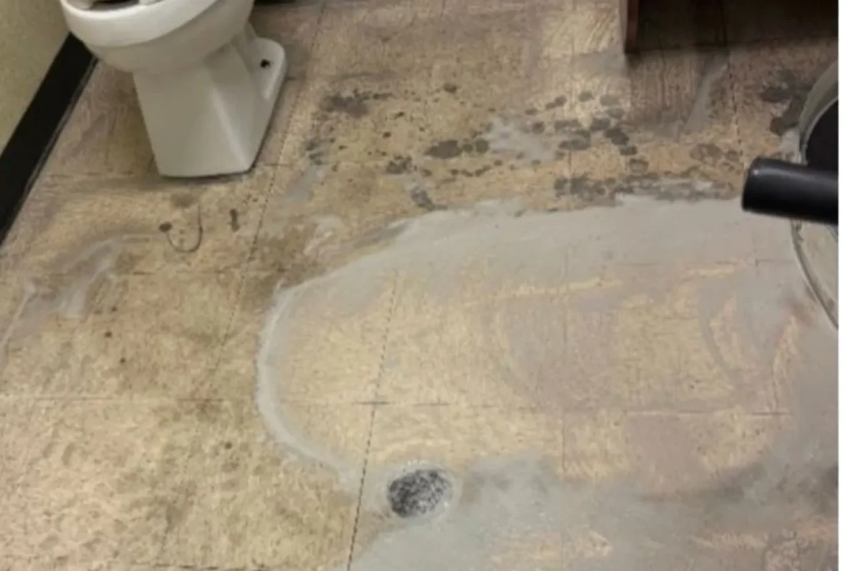 Commercial restroom tile floor heavily stained and wet with grime pooled around floor drain before office cleaning
