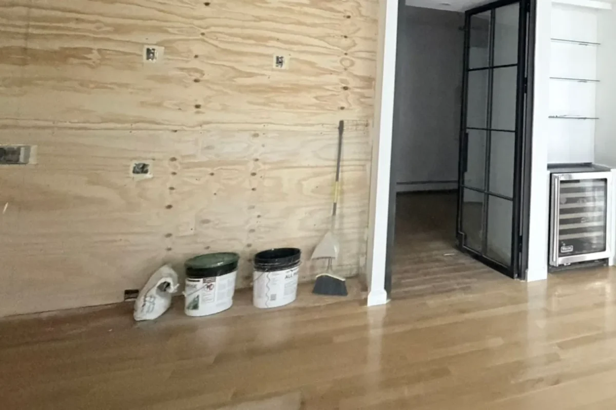 Room mid-renovation with exposed plywood wall, paint buckets and construction materials covering the floor